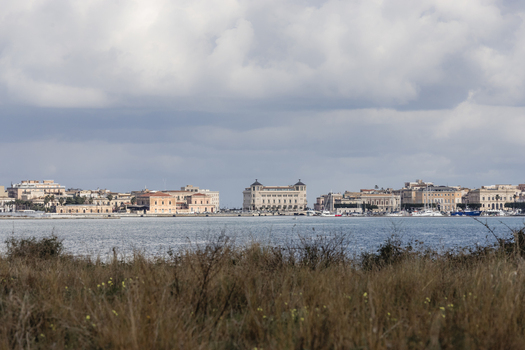View of the historic waterfront of Ortigia across the harbor with traditional architecture under a cloudy sky Syracuse Sicily Italy.