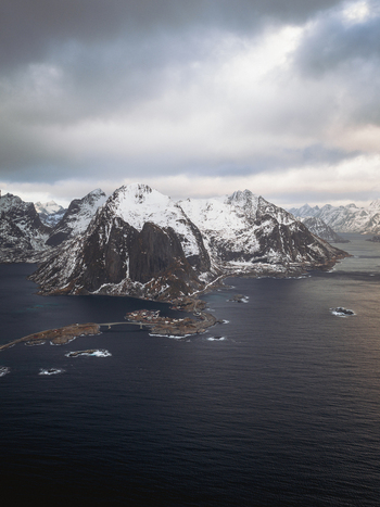 Aerial view of snow capped jagged peaks rise sharply from the dark tranquil waters surrounding Reinebringen Nordland Norway.