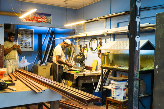 Sri Lanka 20 January 2026 View of two men working in a metal fabrication workshop with aluminum profiles a miter saw and an aquarium against blue walls.