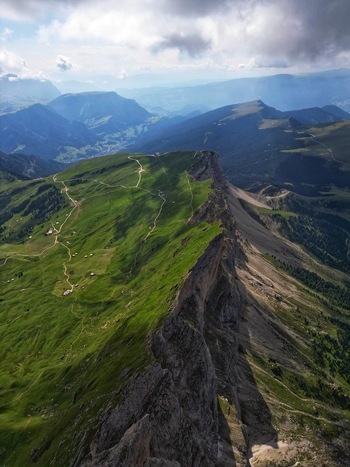 Aerial view of the sharp rocky cliffs and lush green alpine meadows of the Dolomite Mountains with winding hiking trails under a cloudy sky in Canazei Trentino South Tyrol Italy. Aerial view of the sharp rocky cliffs and lush green alpine meadows of the Dolomite Mountains with winding hiking trails under a cloudy sky in Canazei Trentino South Tyrol Italy.