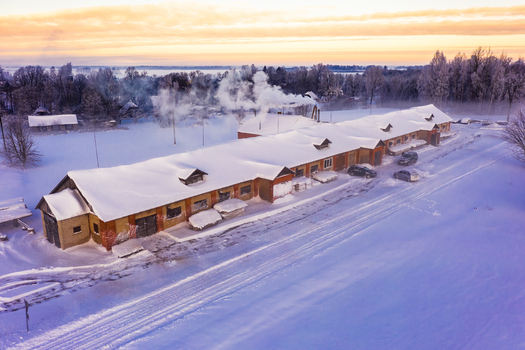 Aerial view of a long brick building covered in snow with smoke rising from a chimney surrounded by a snowy landscape during golden hour in Latvija. Aerial view of a long brick building covered in snow with smoke rising from a chimney surrounded by a snowy landscape during golden hour in Latvija.