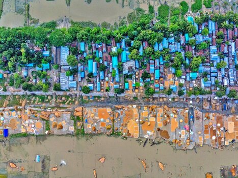Kishoreganj Bangladesh 05 May 2026 Aerial view of a village settlement with blue roofed houses lush green trees and agricultural grain drying areas by the water. Kishoreganj Bangladesh 05 May 2026 Aerial view of a village settlement with blue roofed houses lush green trees and agricultural grain drying areas by the water.