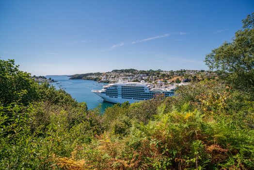 View of a large cruise ship anchored in the Fowey River with the town of Fowey in the background framed by lush trees and ferns under a clear blue sky in Fowey Cornwall United Kingdom. View of a large cruise ship anchored in the Fowey River with the town of Fowey in the background framed by lush trees and ferns under a clear blue sky in Fowey Cornwall United Kingdom.