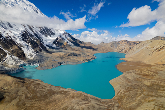 Aerial view of a vibrant turquoise glacial lake nestled among rugged snow capped Himalayan peaks under a bright blue sky in Nepal.