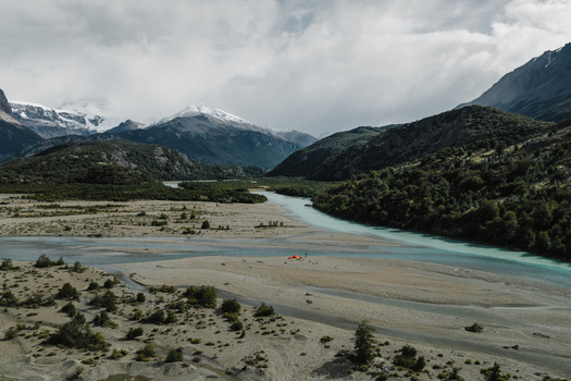 Aerial view of a turquoise glacial river winding through a gravel valley with an orange tent and a person set against snow capped mountains in El Chalten Santa Cruz Province Argentina. Aerial view of a turquoise glacial river winding through a gravel valley with an orange tent and a person set against snow capped mountains in El Chalten Santa Cruz Province Argentina.