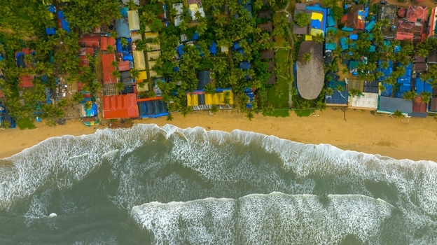 Aerial view of Palolem Beach with colorful beach huts nestled among palm trees along the sandy coastline and gentle waves washing the shore in Canacona Goa India.