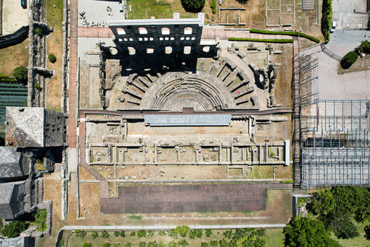 Aerial view of the Roman Theatre of Aosta featuring the semicircular stone seating and the high arched facade wall among ancient ruins in Aosta Aosta Valley Italy. Aerial view of the Roman Theatre of Aosta featuring the semicircular stone seating and the high arched facade wall among ancient ruins in Aosta Aosta Valley Italy.