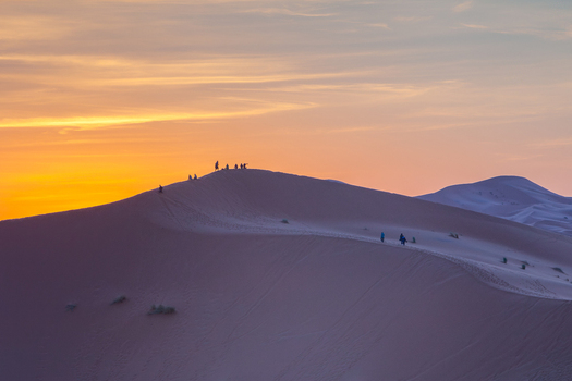 View of people walking along the ridge of a sand dune in the Sahara desert during a vibrant sunset in Morocco.