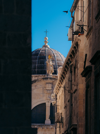 View of the Dubrovnik Cathedral dome and statue framed by narrow stone street walls under a clear blue sky in Dubrovnik Dubrovnik Neretva County Croatia. View of the Dubrovnik Cathedral dome and statue framed by narrow stone street walls under a clear blue sky in Dubrovnik Dubrovnik Neretva County Croatia.