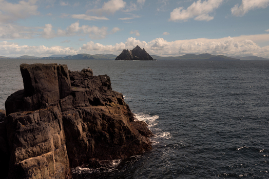 View of Great Skellig rising from the Atlantic Ocean with dark jagged rocks in the foreground and a distant coastline under a cloudy sky County Kerry Ireland. View of Great Skellig rising from the Atlantic Ocean with dark jagged rocks in the foreground and a distant coastline under a cloudy sky County Kerry Ireland.