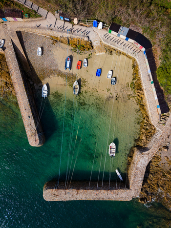 Aerial view of Port Racine with colorful small boats moored in clear turquoise water surrounded by historic stone walls and coastal greenery in La Hague Normandie France.