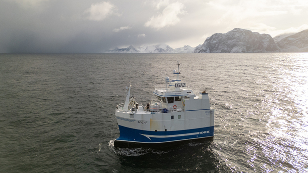 Aerial view of a white and blue fishing boat navigating the cold waters near the snow covered mountains of Reine Nordland Norway.