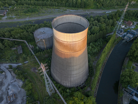 Aerial view of the abandoned cooling tower at the old power plant illuminated by golden hour light surrounded by lush trees and a canal in Charleroi Wallonia Belgium.