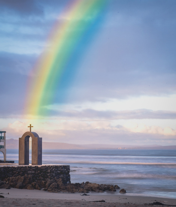 View of a stone arch with a cross on the beach under a vibrant rainbow over the ocean in Langebaan Western Cape South Africa.
