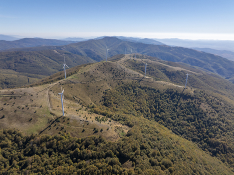 Aerial view of wind turbines situated on a mountain ridge surrounded by dense forest and rolling hills under a clear blue sky in Bulgaria. Aerial view of wind turbines situated on a mountain ridge surrounded by dense forest and rolling hills under a clear blue sky in Bulgaria.