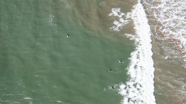 Aerial view of surfers waiting for waves in the green sea with white foam near the shore at Bantham Beach in Kingsbridge Devon United Kingdom. Aerial view of surfers waiting for waves in the green sea with white foam near the shore at Bantham Beach in Kingsbridge Devon United Kingdom.
