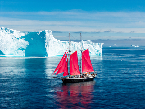 Aerial view of a sailboat with vibrant red sails navigating the deep blue waters near a massive white iceberg under a clear sky in Greenland. Aerial view of a sailboat with vibrant red sails navigating the deep blue waters near a massive white iceberg under a clear sky in Greenland.
