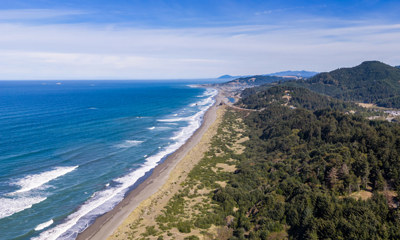 Aerial view of the rugged coastline with waves crashing on the sandy beach dense evergreen forests and rolling hills under a clear blue sky in Gold Beach Oregon United States. Aerial view of the rugged coastline with waves crashing on the sandy beach dense evergreen forests and rolling hills under a clear blue sky in Gold Beach Oregon United States.