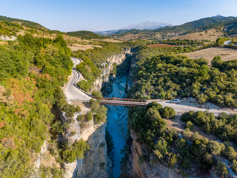 Aerial view of Osumi canyon featuring a metal bridge crossing the river surrounded by lush green forests rocky cliffs and a winding road in Dhores Berat County Albania.