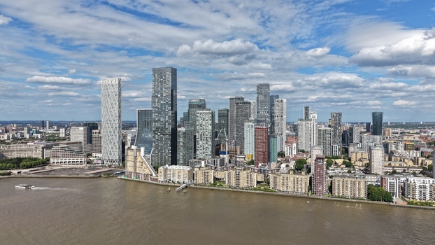 London United Kingdom 16 June 2026 Aerial view of Canary Wharf skyscrapers and modern office buildings along the River Thames under a cloudy blue sky.