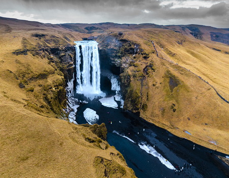Aerial view of the majestic Skógafoss waterfall cascading down rugged cliffs into a dark river contrasting with the golden arid landscape Skógafoss Rangárþing eystra Iceland. Aerial view of the majestic Skógafoss waterfall cascading down rugged cliffs into a dark river contrasting with the golden arid landscape Skógafoss Rangárþing eystra Iceland.