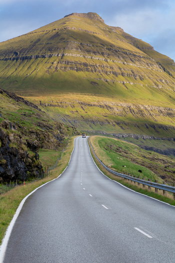 View of a winding road leading through a dramatic mountainous landscape with green slopes and rocky terrain under a cloudy sky in Faroe Islands. View of a winding road leading through a dramatic mountainous landscape with green slopes and rocky terrain under a cloudy sky in Faroe Islands.