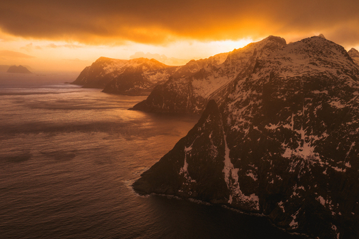 Aerial view of jagged snow dusted peaks meet the turbulent sea under a fiery sunset sky Lofoten Nordland Norway.