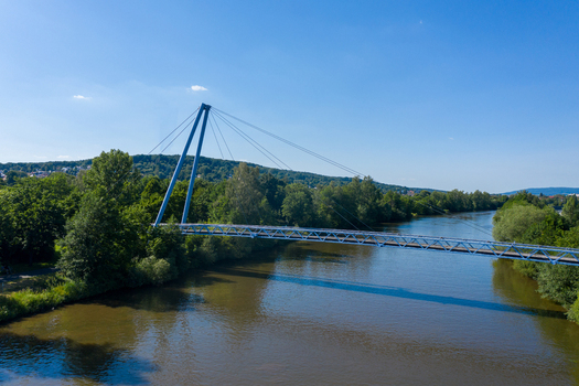 Aerial view of the Lila Brucke bridge crossing the Regnitz river surrounded by lush green trees and forested hills under a clear blue sky in Forchheim Bavaria Germany. Aerial view of the Lila Brucke bridge crossing the Regnitz river surrounded by lush green trees and forested hills under a clear blue sky in Forchheim Bavaria Germany.