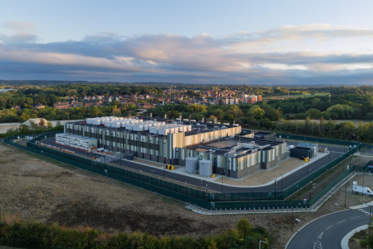 Aerial view of a modern data center facility surrounded by a secure perimeter fence industrial cooling units on the roof and lush green trees in the United Kingdom. Aerial view of a modern data center facility surrounded by a secure perimeter fence industrial cooling units on the roof and lush green trees in the United Kingdom.