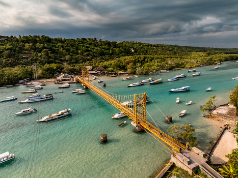 Aerial view of the iconic yellow suspension bridge connecting Nusa Lembongan and Nusa Ceningan surrounded by turquoise waters boats and lush green vegetation in Nusa Penida Bali Indonesia. Aerial view of the iconic yellow suspension bridge connecting Nusa Lembongan and Nusa Ceningan surrounded by turquoise waters boats and lush green vegetation in Nusa Penida Bali Indonesia.