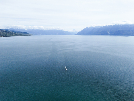 Aerial view of a lone sailboat on the vast blue waters of Lake Geneva with distant mountains under a cloudy sky in Lausanne Vaud Switzerland.