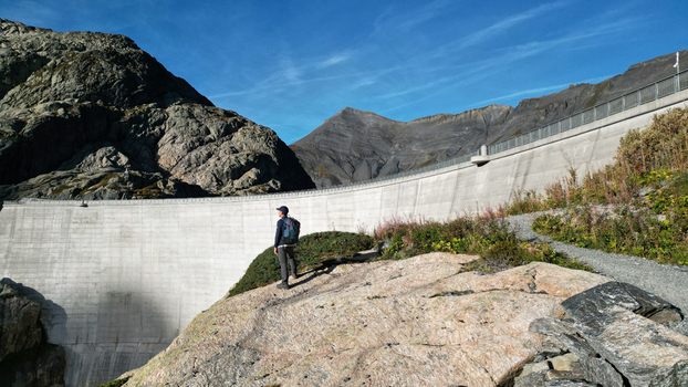 Aerial view of a hiker on a rocky ledge overlooking the massive concrete wall of the Vieux Emosson Dam set against rugged alpine mountains in Finhaut Valais Switzerland. Aerial view of a hiker on a rocky ledge overlooking the massive concrete wall of the Vieux Emosson Dam set against rugged alpine mountains in Finhaut Valais Switzerland.