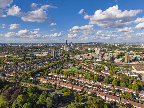Aerial view of the residential neighborhood with rows of houses lush green trees and a distant city skyline under a bright blue sky in Brentford England United Kingdom. Aerial view of the residential neighborhood with rows of houses lush green trees and a distant city skyline under a bright blue sky in Brentford England United Kingdom.