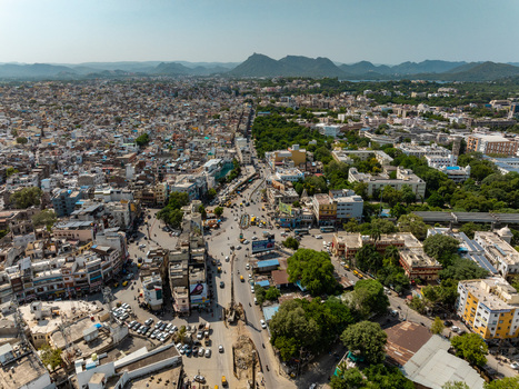 Aerial view of the dense urban landscape of Udaipur featuring busy streets traditional buildings lush green trees and distant hills under a clear sky in Udaipur Rajasthan India.