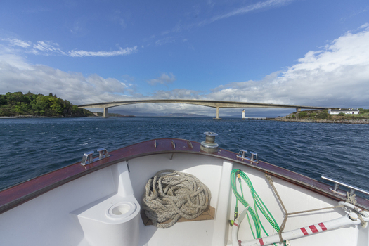 View of Skye Bridge seen from the bow of a boat on the water with green hills and a blue sky in Isle of Skye United Kingdom. View of Skye Bridge seen from the bow of a boat on the water with green hills and a blue sky in Isle of Skye United Kingdom.
