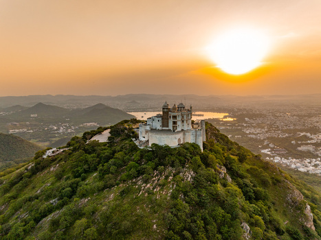 Aerial view of the historic Monsoon Palace on a hilltop overlooking Udaipur surrounded by lush green hills and a distant lake under a golden sunset sky in Udaipur Rajasthan India.