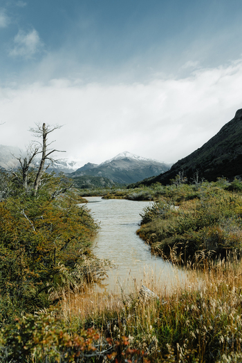 View of the river flowing through the rugged landscape with snow capped mountains in the background and lush vegetation along the banks in El Chalten Santa Cruz Province Argentina. View of the river flowing through the rugged landscape with snow capped mountains in the background and lush vegetation along the banks in El Chalten Santa Cruz Province Argentina.