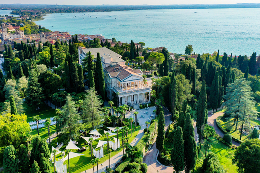 Aerial view of Villa Cortine surrounded by lush cypress trees and green gardens with Lake Garda in the background in Sirmione Lombardia Italy. Aerial view of Villa Cortine surrounded by lush cypress trees and green gardens with Lake Garda in the background in Sirmione Lombardia Italy.