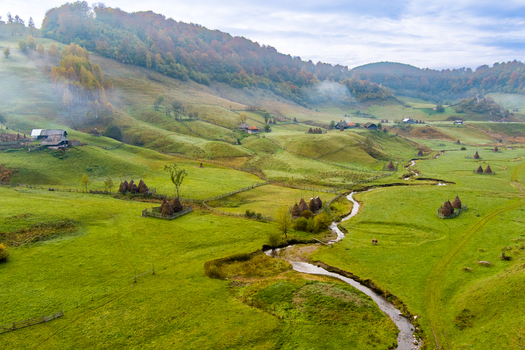 Aerial view of Fundatura Ponorului featuring a winding stream through green meadows traditional haystacks and rolling hills under morning mist in Ohaba Ponor Hunedoara Romania.