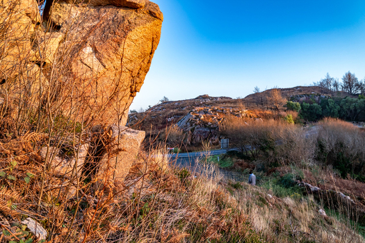 View of Kincasslagh Mass Rock surrounded by rugged terrain dry vegetation and a winding road under a clear blue sky during golden hour in Kincasslagh County Donegal Ireland. View of Kincasslagh Mass Rock surrounded by rugged terrain dry vegetation and a winding road under a clear blue sky during golden hour in Kincasslagh County Donegal Ireland.