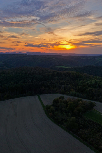 Ehlscheid Rhineland Palatinate Germany 26 September 2026 Aerial view of rolling hills and forests at sunset with agricultural fields under a vibrant orange sky.