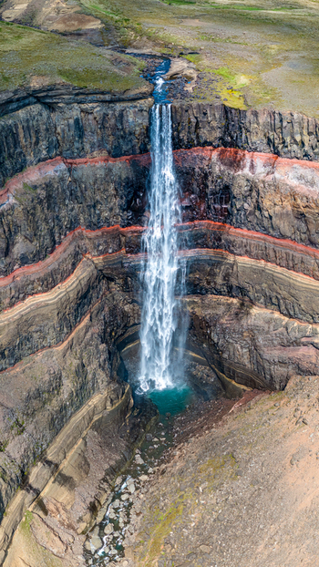Aerial view of the majestic Hengifoss waterfall cascading down a deep canyon with distinct red clay layers embedded in the basalt rock strata Iceland. Aerial view of the majestic Hengifoss waterfall cascading down a deep canyon with distinct red clay layers embedded in the basalt rock strata Iceland.