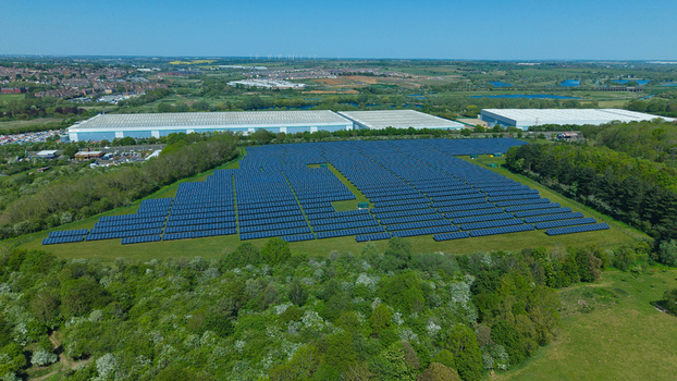 Aerial view of a large solar farm array situated in a green field surrounded by lush trees and industrial warehouses under a clear blue sky in Little Irchester England United Kingdom. Aerial view of a large solar farm array situated in a green field surrounded by lush trees and industrial warehouses under a clear blue sky in Little Irchester England United Kingdom.