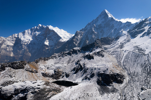 Aerial view of the snow capped Himalayan peaks rugged rocky slopes and a small dark glacial lake under a clear blue sky in Nepal.