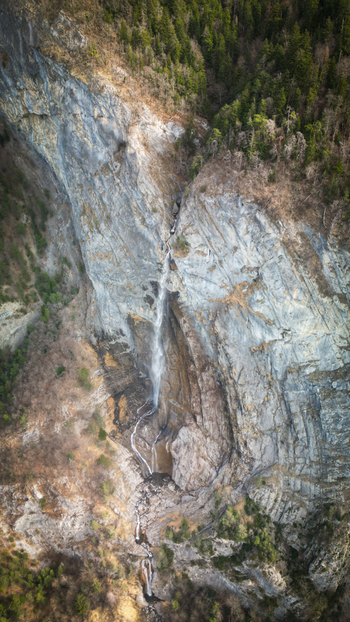 Aerial view of a delicate waterfall cascading down a rugged rock face its white water contrasting with the gray stone and the green trees Sallanches Auvergne Rhône Alpes France. Aerial view of a delicate waterfall cascading down a rugged rock face its white water contrasting with the gray stone and the green trees Sallanches Auvergne Rhône Alpes France.