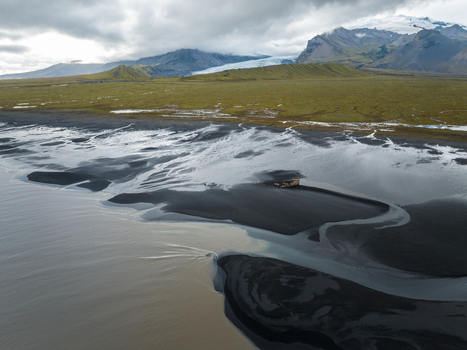 Aerial view of a stark black sand beach meeting the glacial waters under an overcast sky with mountains rising in the distance Skaftafell Iceland. Aerial view of a stark black sand beach meeting the glacial waters under an overcast sky with mountains rising in the distance Skaftafell Iceland.