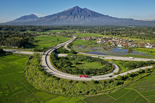 Aerial view of the Jalan Tol Bawen Salatiga highway curving through lush green rice fields with Mount Merbabu in the background Kecamatan Tengaran Jawa Tengah Indonesia.