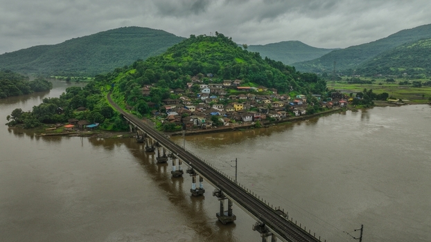 Aerial view of a railway bridge crossing a river with a small village nestled at the base of a lush green hill under a cloudy sky in Mahad Maharashtra India.