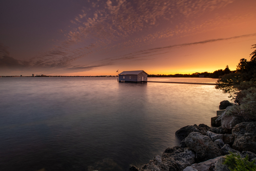 View of the iconic Blue Boat House standing on the Swan River at sunset with calm water rocky shoreline and a vibrant orange sky in Crawley Western Australia Australia. View of the iconic Blue Boat House standing on the Swan River at sunset with calm water rocky shoreline and a vibrant orange sky in Crawley Western Australia Australia.