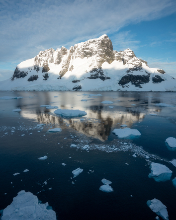 View of a massive snow covered mountain peak reflecting in the calm dark blue waters filled with floating icebergs and ice floes under a clear sky in Antarctica.